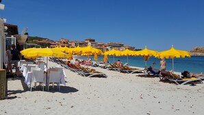 Een strand met rijen ligbedden en gele parasols, een helderblauwe lucht en gebouwen op de achtergrond.