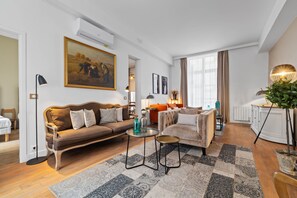 Ein Wohnzimmer mit braunem Sofa, einem runden Couchtisch und einem beige gestreiften Sessel. An der Wand hängt ein Gemälde, eine Zimmerpflanze steht bereit und ein Fenster ist mit Vorhängen versehen.