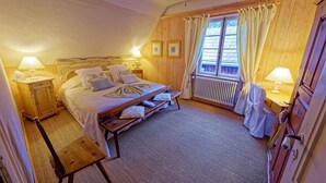 Un dormitorio acogedor con cabecera de madera, una cama grande con almohadas, un banco al pie de la cama, un radiador y una ventana con cortinas.