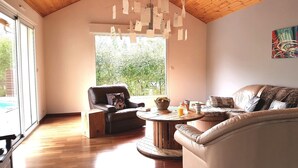 Amplia sala de estar con techo alto, un ventanal que ofrece vistas a la vegetación, un sofá de cuero, un sillón de cuero y una mesa de centro de madera con un cesto de pan y un vaso de jugo de naranja.