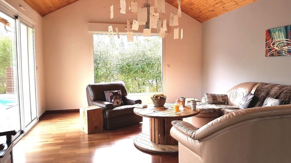 A spacious living room with a high ceiling, a large window offering a view of greenery, a leather sofa, a leather armchair, and a wooden coffee table with a basket of bread and a glass of orange juice.