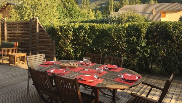 Un área de comedor al aire libre con una mesa preparada para cuatro personas, rodeada de sillas de madera y un deck de madera.