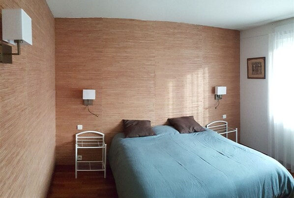 Een slaapkamer met een houten wand, twee nachtkastjes, een bed met een blauwe deken, twee wandlampen en een raam met gordijnen.