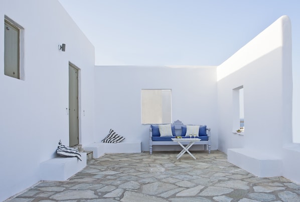 A white-walled room with a stone floor, a blue sofa with cushions, a small table, and a window with a white blind.