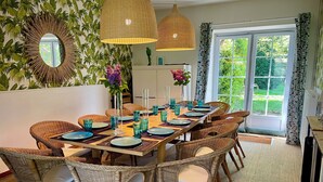 Un coin repas avec des chaises en osier, une table en bois dressée avec des assiettes et des verres, et un grand miroir orné d’un cadre en forme de soleil.