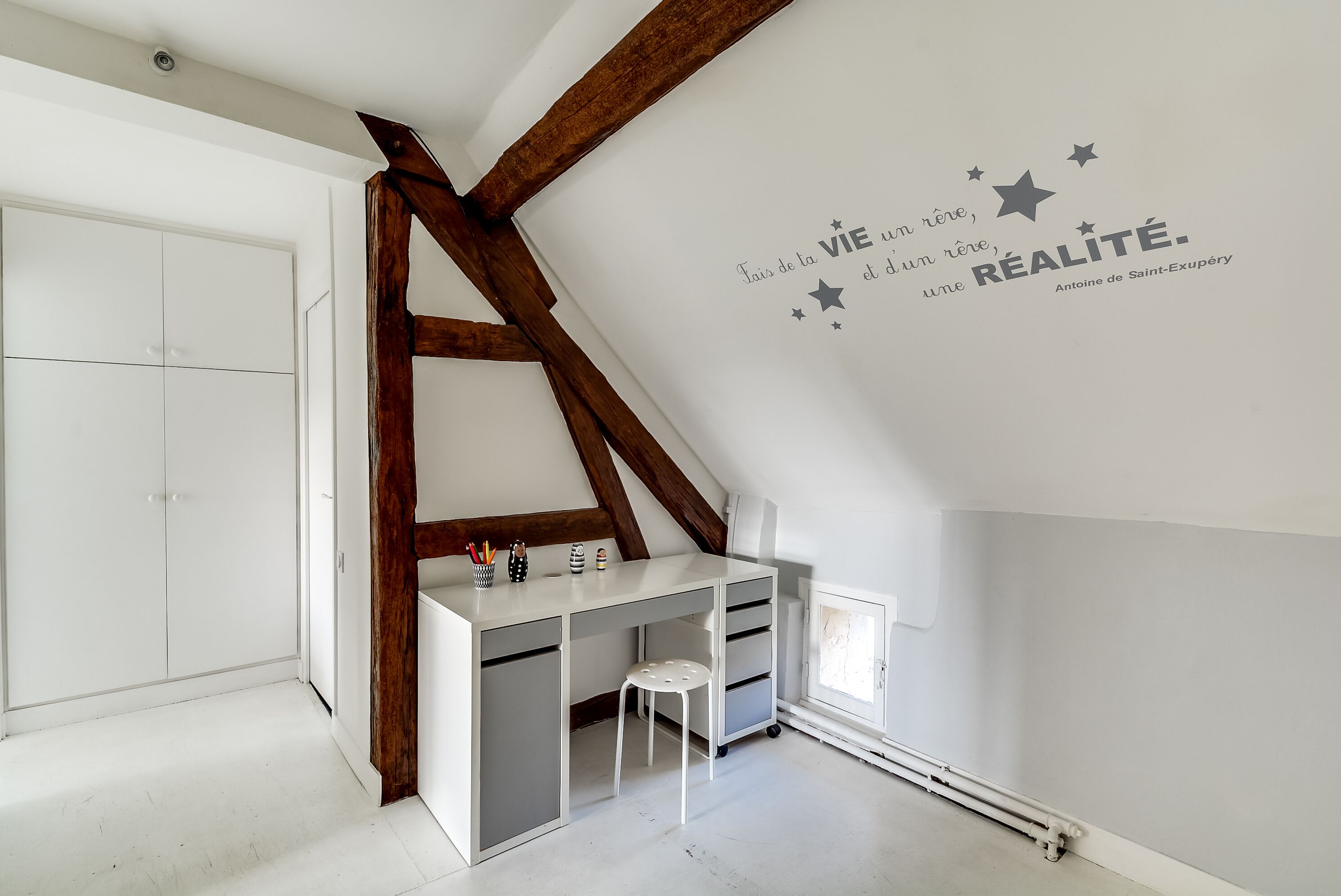 Una habitación con techo inclinado, un escritorio blanco con cajones, una silla y un adhesivo de pared con estrellas y texto.