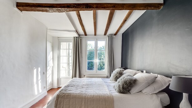 A bedroom with a large bed, a window with curtains, a radiator, and a wooden ceiling beam.