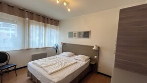 Ein Schlafzimmer mit Bett, Stuhl, einem Fenster mit Vorhängen, einem Wandventilator und einer Deckenleuchte.