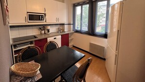 Eine kompakte Küche mit weißen Schränken, einer Mikrowelle, einer Kaffeemaschine und einem Kühlschrank. Ein Esstisch mit Stühlen und ein Fenster mit Vorhängen sind ebenfalls vorhanden.