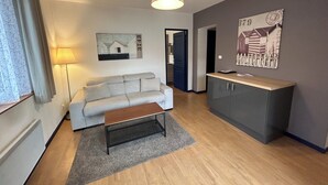 Ein modernes Wohnzimmer mit einem grauen Sofa, einem Holztisch und einem grauen Teppich. Eine Lampe, ein Gemälde und ein Schrank mit Arbeitsplatte sind ebenfalls zu sehen.