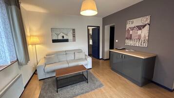 Ein modernes Wohnzimmer mit einem grauen Sofa, einem Holztisch und einem grauen Teppich. Eine Lampe, ein Gemälde und ein Schrank mit Arbeitsplatte sind ebenfalls zu sehen.