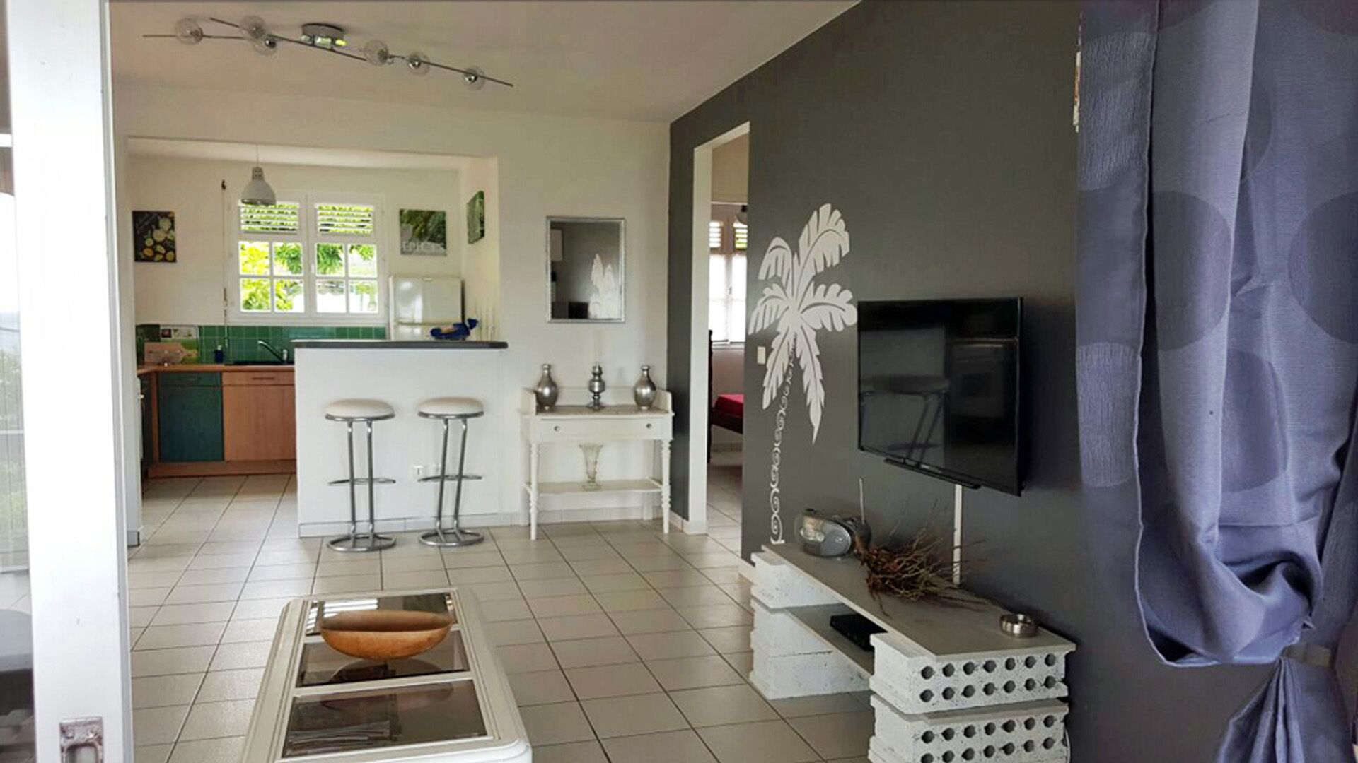 Eine moderne Küche mit weißen Schränken, einer zentralen Kochinsel mit Hockern, einem Essbereich mit weißem Tisch und einem an einer dunklen Wand montierten Fernseher.