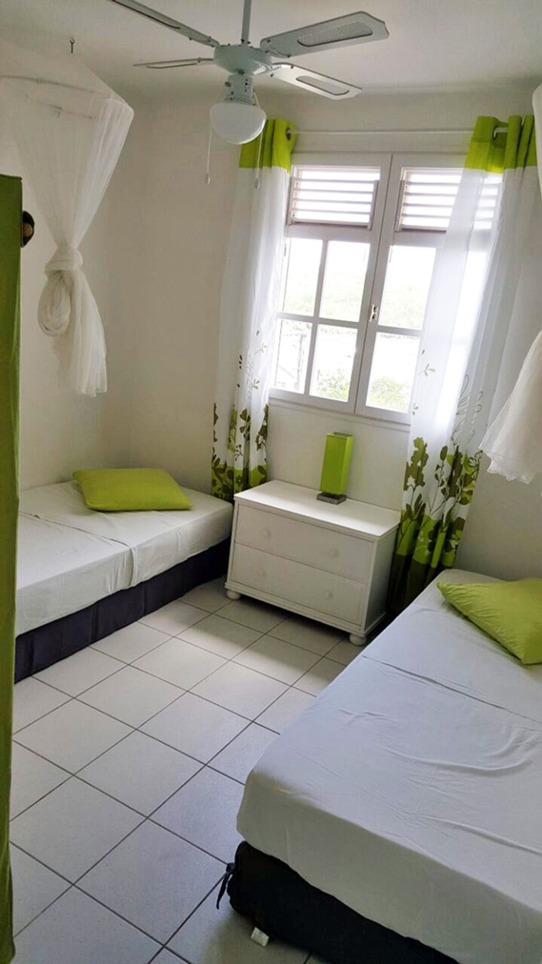 Ein kleiner, sauberer Raum mit zwei Betten, einem Nachttisch, einem Fenster mit Vorhängen und einem Deckenventilator.
