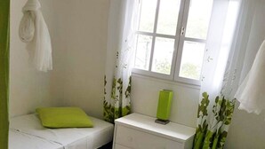Une petite chambre propre, avec deux lits, une table de chevet, une fenêtre avec des rideaux et un ventilateur de plafond.
