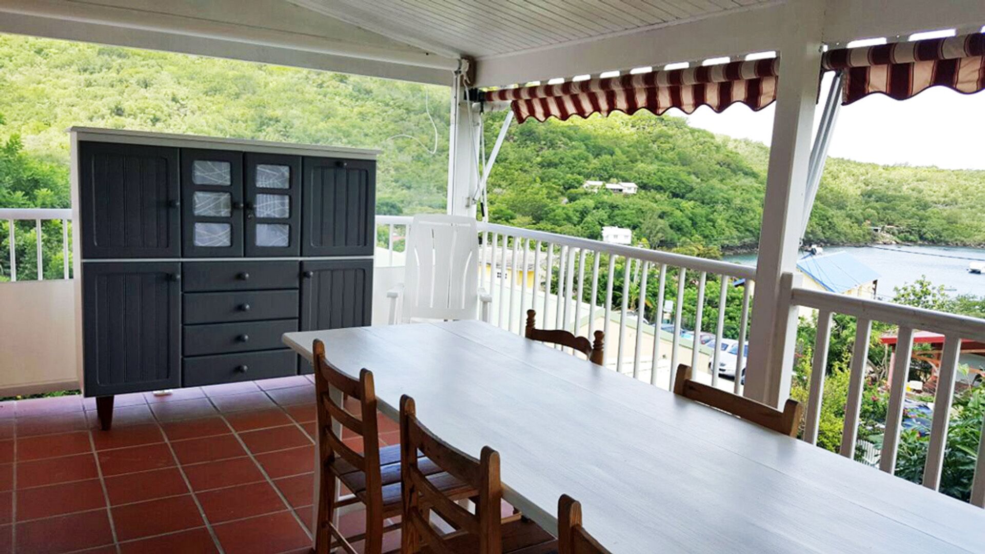 A balcony with a white table and chairs, a grey cabinet, and a view of a lush green landscape and water.