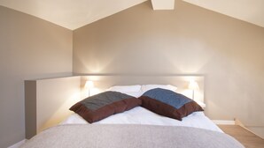 Ein Schlafzimmer mit schrägen Wänden, einem Bett mit weißer Bettwäsche, zwei Kissen und zwei Nachttischlampen.