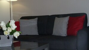 Un sofá negro con cojines estampados en rojo, negro y blanco, una mesa de centro de vidrio con un jarrón de flores blancas y una lámpara de pie.