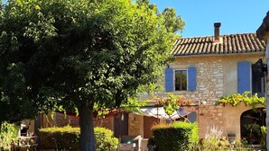Una casa de piedra con techo de tejas, un árbol grande en primer plano y un patio con mobiliario exterior.