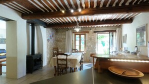 Un interior rústico con pared de piedra, vigas de madera en el techo, un comedor con sillas y una mesa, y una zona de cocina con estufa.