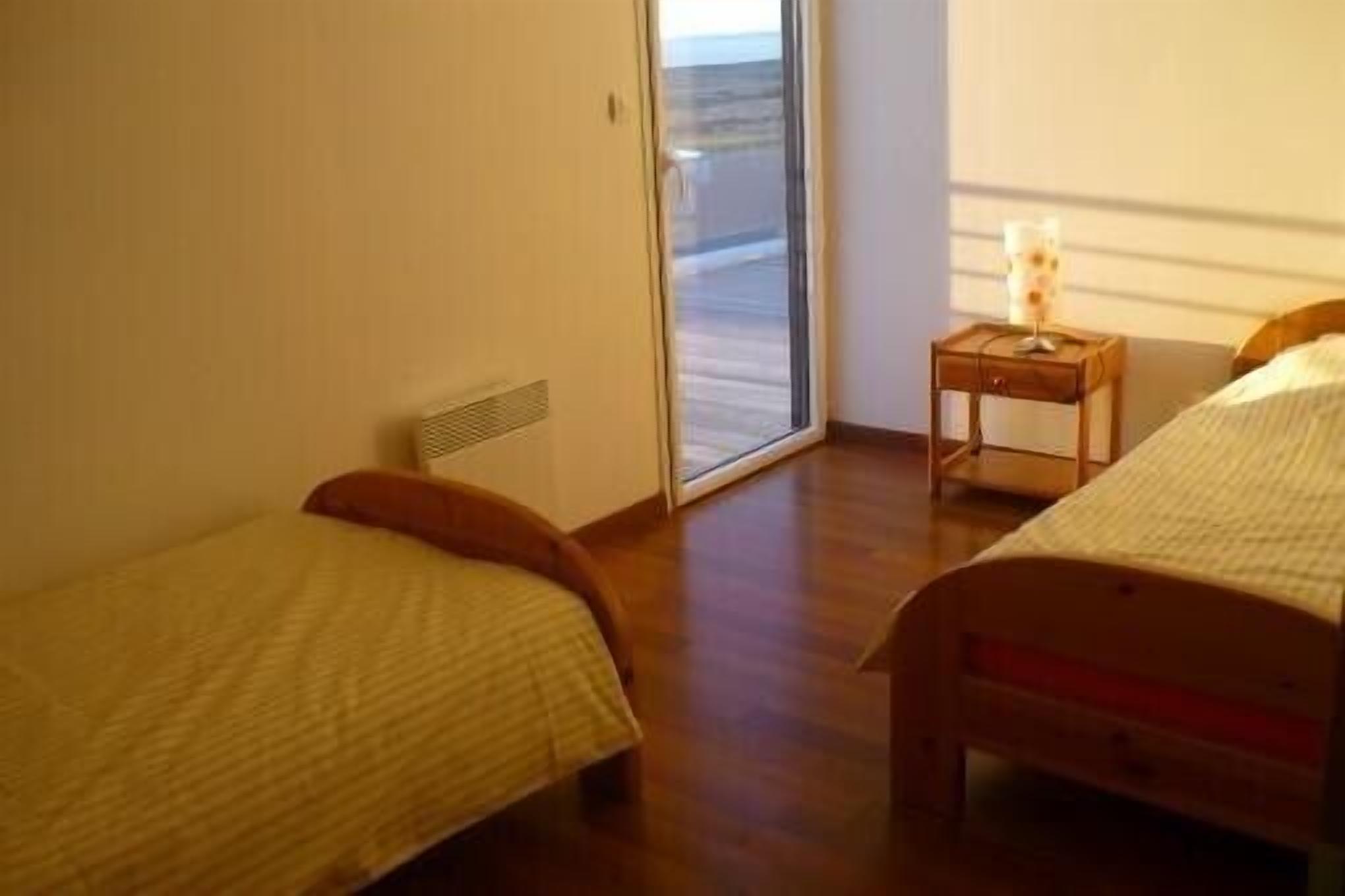 Quarto com duas camas de solteiro, uma pequena mesa de cabeceira com abajur e vista por uma porta de vidro.