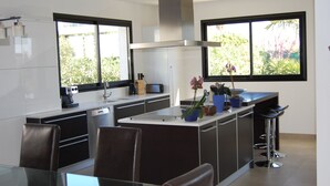 A modern kitchen with a central island, bar stools, and a dining area with a glass table. - Direct access to the beach, swimming pool, 8 people, modern 5-star villa (Sète)