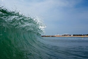 A large wave in the ocean with a distant view of a coastal area including a building and a pier.