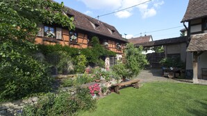 Une maison traditionnelle à colombages, avec des fondations en pierre, un banc en bois et un jardin bien entretenu où poussent diverses plantes et fleurs.