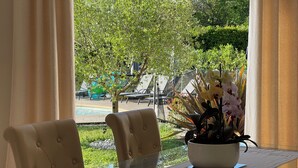 Un coin repas avec une table en verre et des chaises rembourrées, un pot d’orchidée et une vue sur une piscine et la verdure.