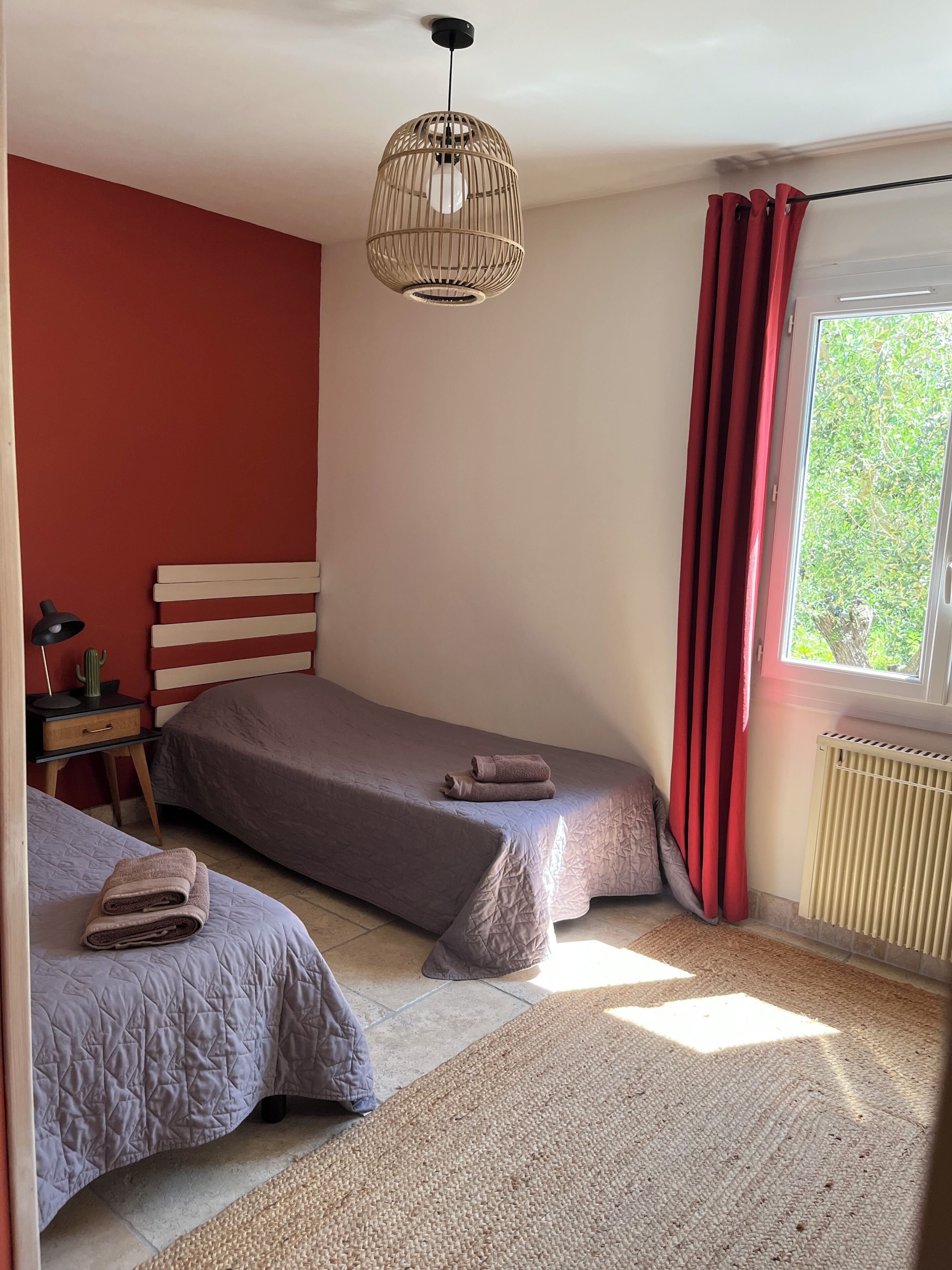 A bedroom with two beds, a bedside table with a lamp, a hanging light fixture, a window with curtains, and a radiator.