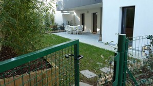 En moderne bygning med en terrasse, møbleret med et bord og stole, omgivet af grønne planter og en indhegnet have.