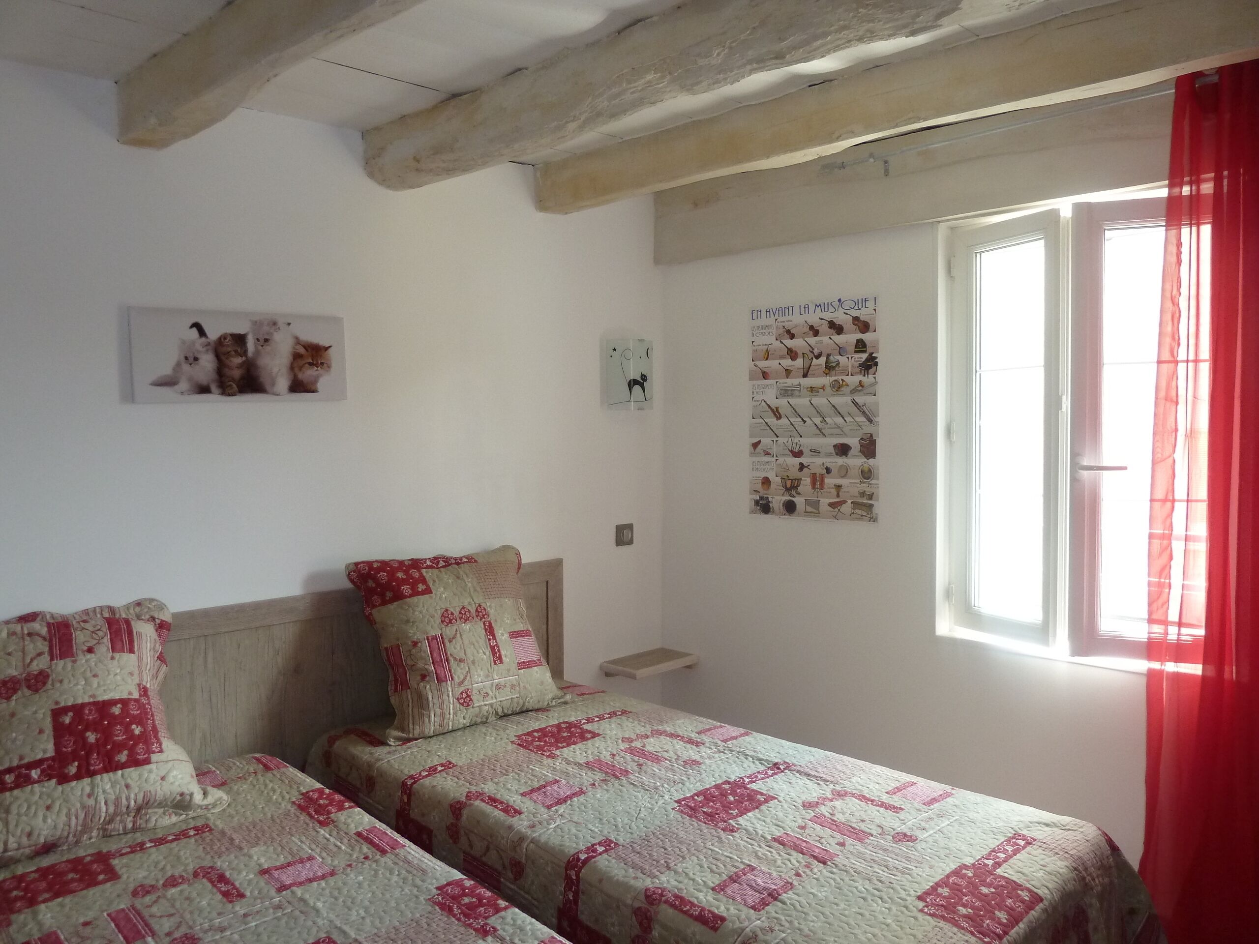 A bedroom with two beds, a window with red curtains, a wall poster, and a small shelf.