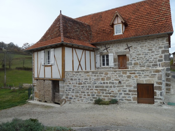 Une maison en pierre à deux étages, avec un toit recouvert de tuiles rouges, des poutres en bois et une petite fenêtre.