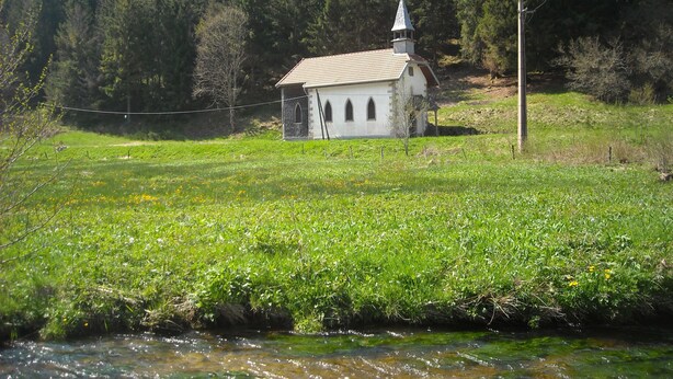 En lille hvid kirke med et klokketårn, omgivet af frodig vegetation og en klar å.
