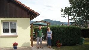 Et hus med en overdækket terrasse, et vindue med blondegardiner og en potteplante med røde blomster.