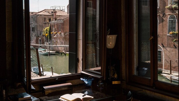 Quarto com uma grande janela com vista para um canal, com uma mesa e cadeiras, e vista para edifícios históricos.