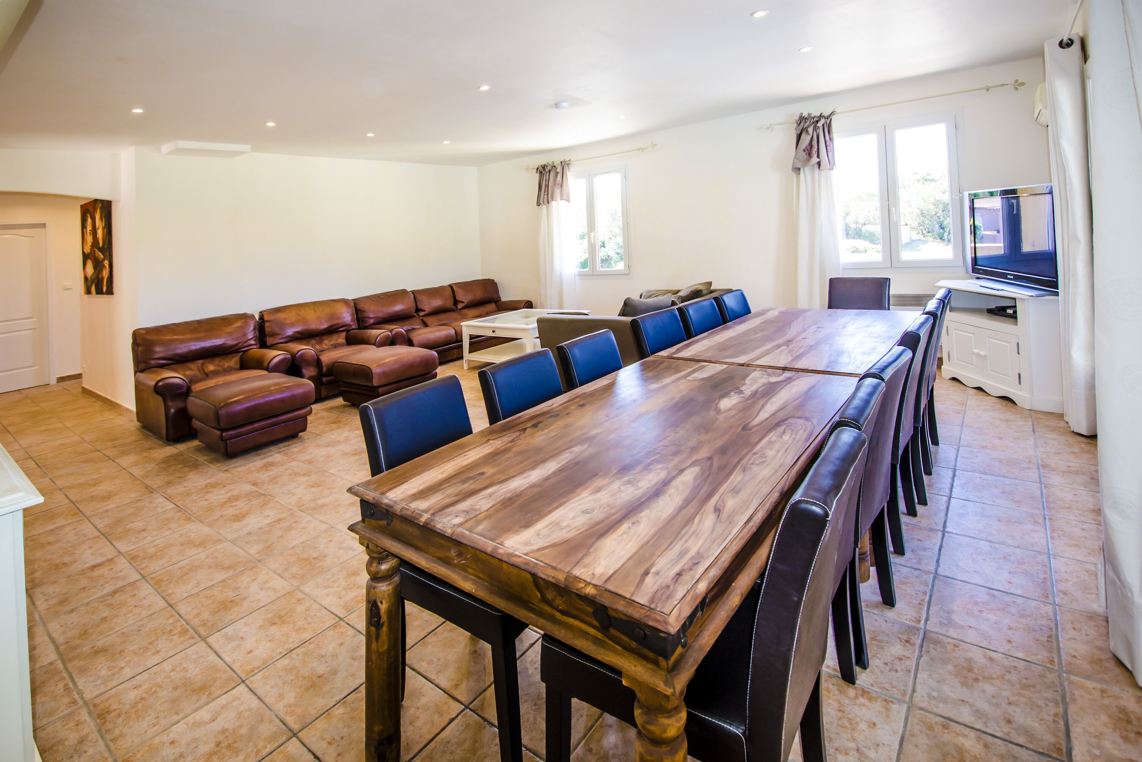 A spacious living room with a long wooden dining table, blue chairs, a brown leather sofa, and a television on a stand.