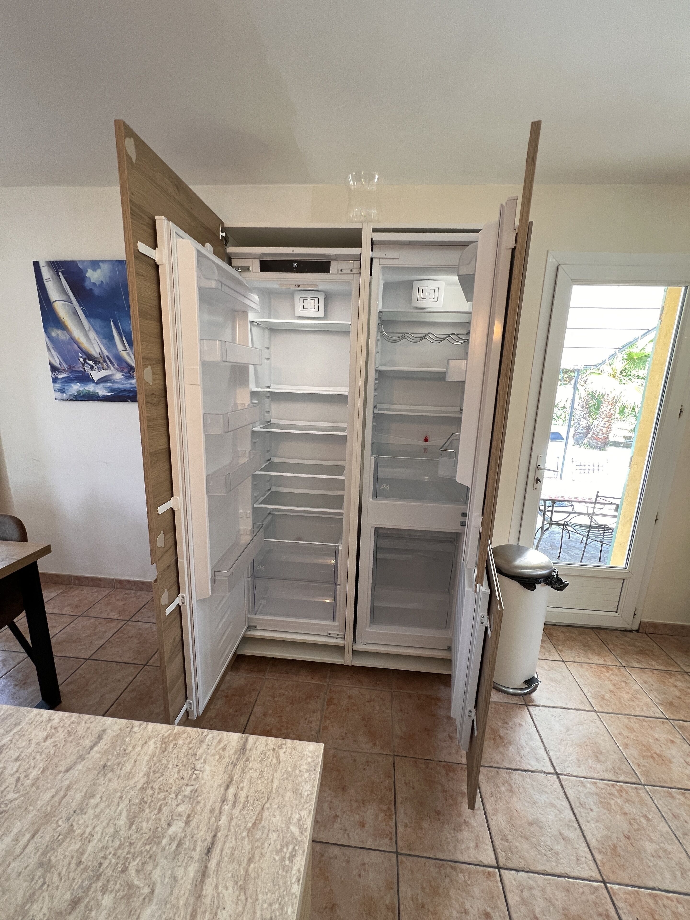Una cocina con un refrigerador abierto, un cesto de basura y una mesa de comedor.