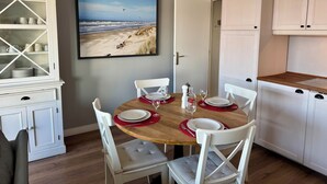 Et spiseområde med et træbord dækket til fire personer, hvide stole, et hvidt skab med glasdøre, og et billede af en strandscene på væggen.