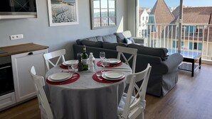 En moderne stue med spiseplads, en grå sofa, et spisebord dækket med tallerkener og glas, samt et stort vindue med udsigt til bygninger.