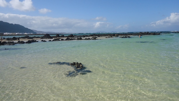 Una playa con agua turquesa cristalina, afloramientos rocosos y montañas a lo lejos.