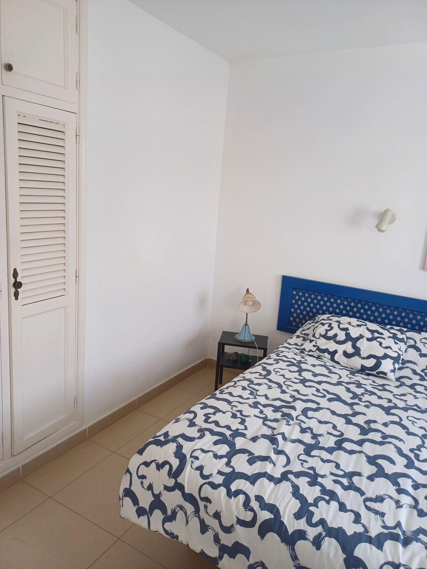 Een slaapkamer met een bed, nachtkastje, lamp, garderobe en een blauw hoofdeinde.