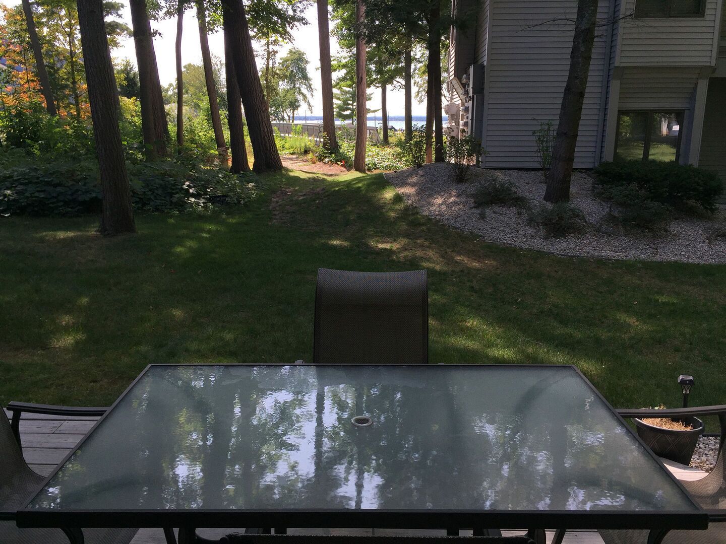 A patio with a glass-top table and chairs, surrounded by a grassy area with trees and a view of a body of water.