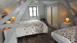 A bedroom with a sloped ceiling, a window with curtains, a bed with a geometric-patterned bedspread, a bedside table with a lamp, and a chair. - House made of regional stones, typical from Auvergne, greenery landscape (Gouttières)
