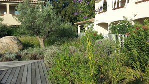 A wooden deck leading to a garden with various plants and flowers, a large rock, and a building with balconies and a white fence. - Modern mini villa T3 800 m from the beach - all comforts, air conditioning, terrace (Lecci)