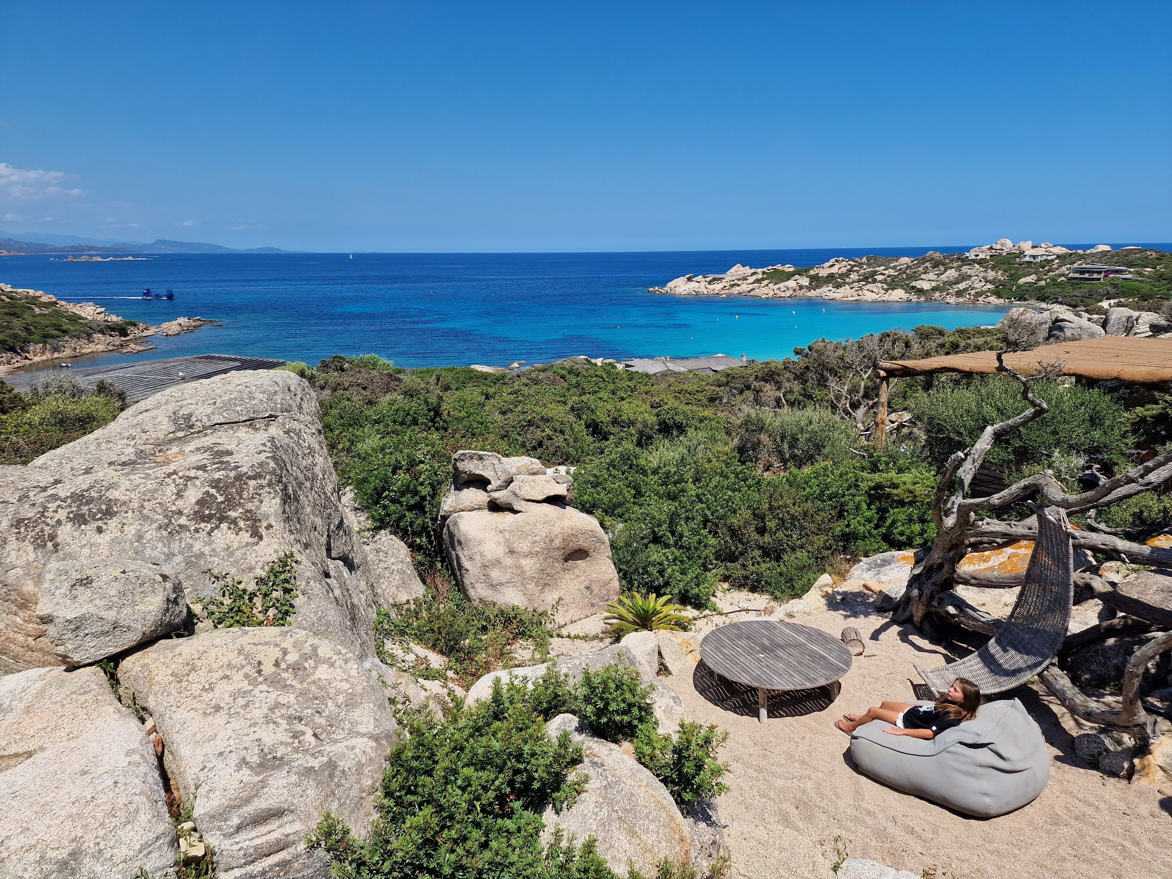 Eine Küstenlandschaft mit felsigem Gelände, blauem Meer und einer kleinen Strandbucht mit einer Holzterrasse und Liegestühlen.
