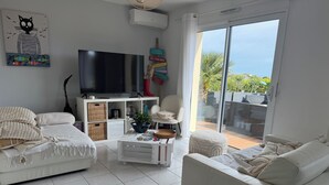A modern living room with a flat-screen TV, white sofas, a coffee table, and a sliding glass door leading to an outdoor area.