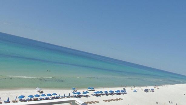 A beach with white sand and clear turquoise water. There are blue umbrellas and lounge chairs along the shore.