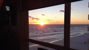 A room with a large window overlooking a beach at sunset.