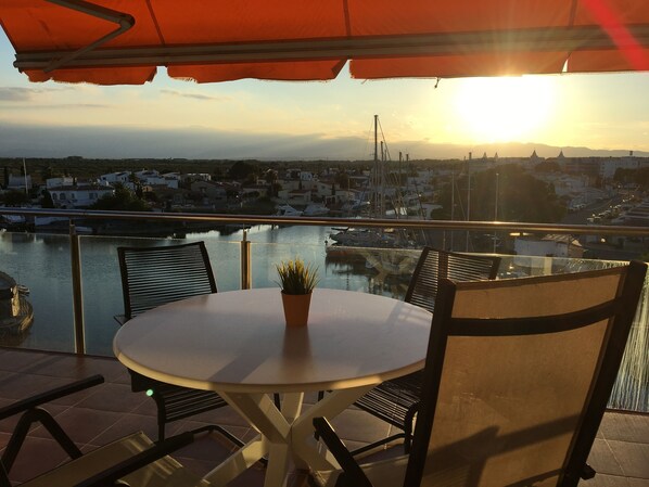 A table with chairs and a potted plant on a balcony overlooking a marina and cityscape during sunset. - Panoramic view of the Mediterrannée, apartment on canal near beach  (Rosas)