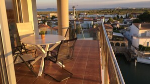 A balcony with a table and chairs, overlooking a coastal town with buildings and a lighthouse in the distance. - Panoramic view of the Mediterrannée, apartment on canal near beach  (Rosas)